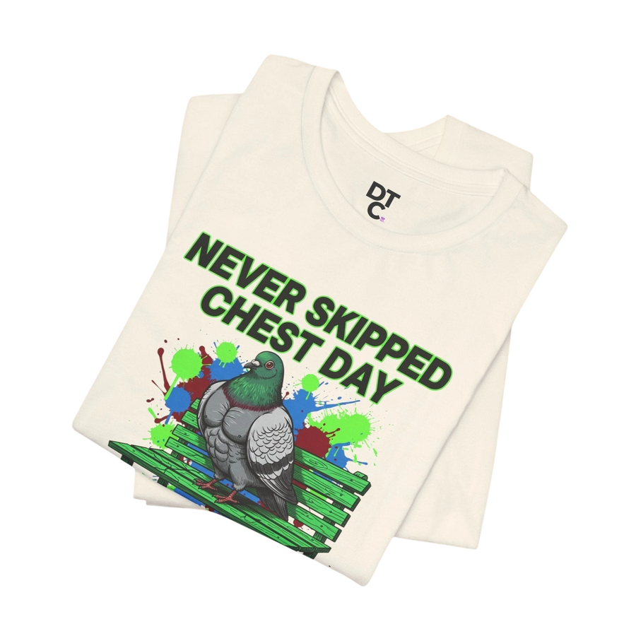 DailyThreadCo. cream cotton t-shirt with a graphic of a muscular pigeon on a green bench, surrounded by colorful paint splatters and the text "NEVER SKIPPED CHEST DAY."