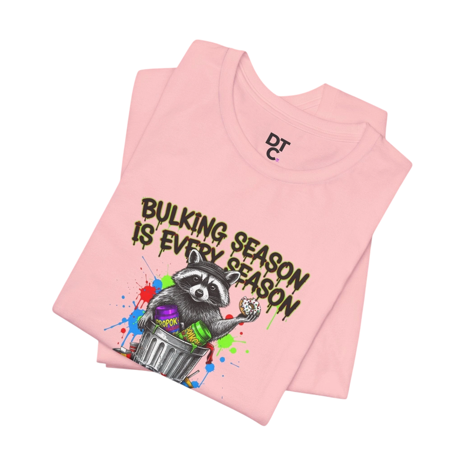 DailyThreadCo. pink 100% cotton t-shirt with a graphic of a raccoon in a trash can holding a donut and the text "BULKING SEASON IS EVERY SEASON".