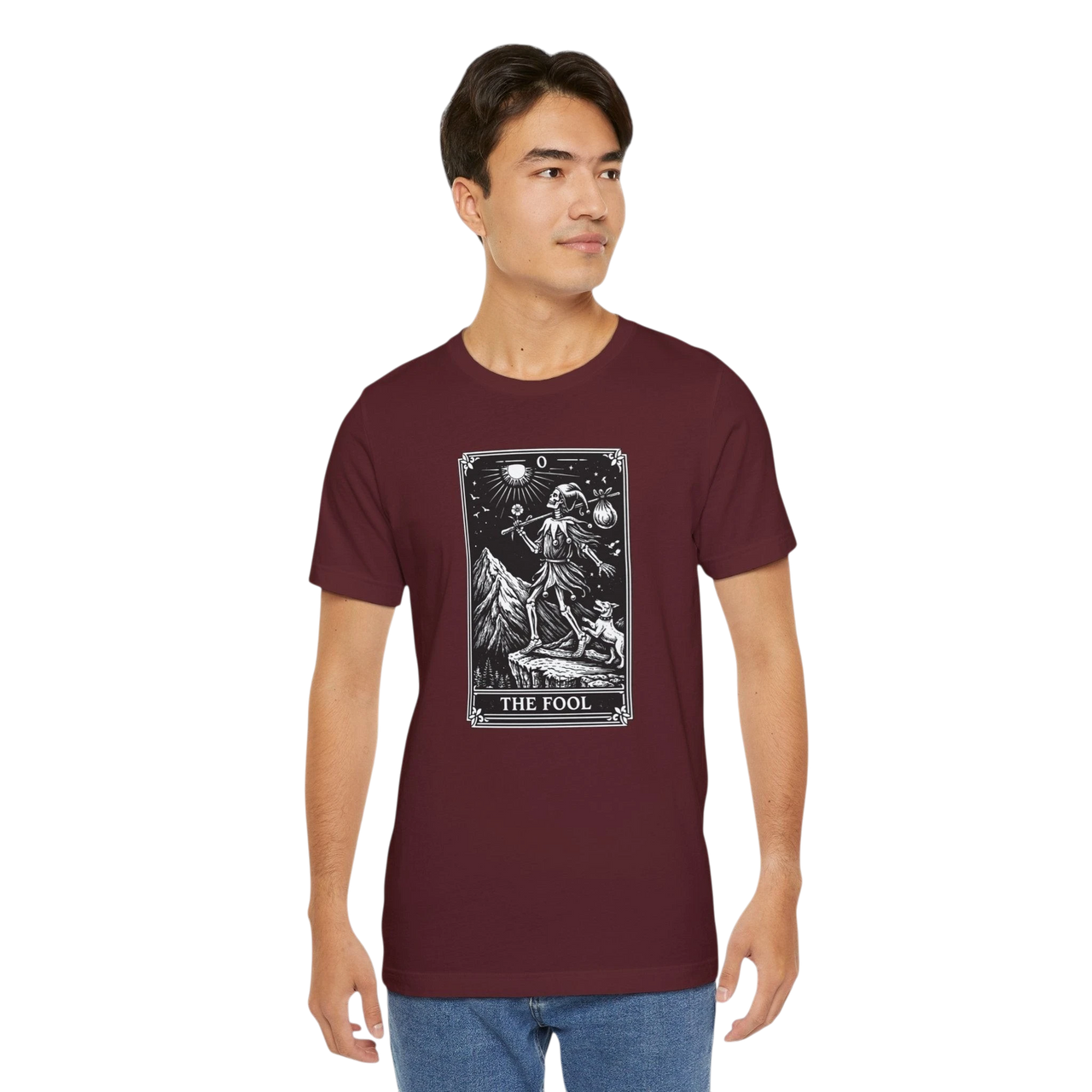 DailyThreadCo. maroon cotton t-shirt with 'The Fool' tarot card graphic print on the front. Features a skeleton figure on a mountain cliff with a dog.