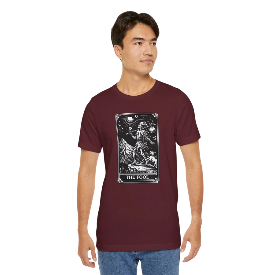 DailyThreadCo. maroon cotton t-shirt with 'The Fool' tarot card graphic print on the front. Features a skeleton figure on a mountain cliff with a dog.