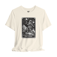 DailyThreadCo. cream cotton t-shirt with 'The Fool' tarot card graphic in black and white. Features a skeleton figure, dog, mountains, and sun.