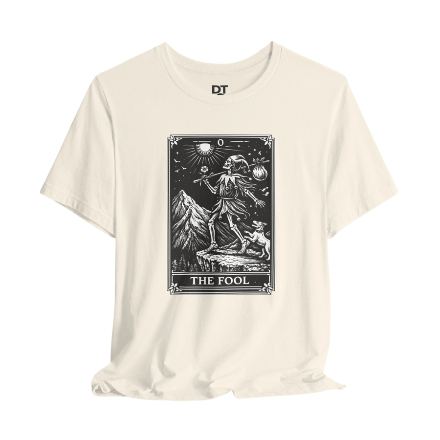 DailyThreadCo. cream cotton t-shirt with 'The Fool' tarot card graphic in black and white. Features a skeleton figure, dog, mountains, and sun.