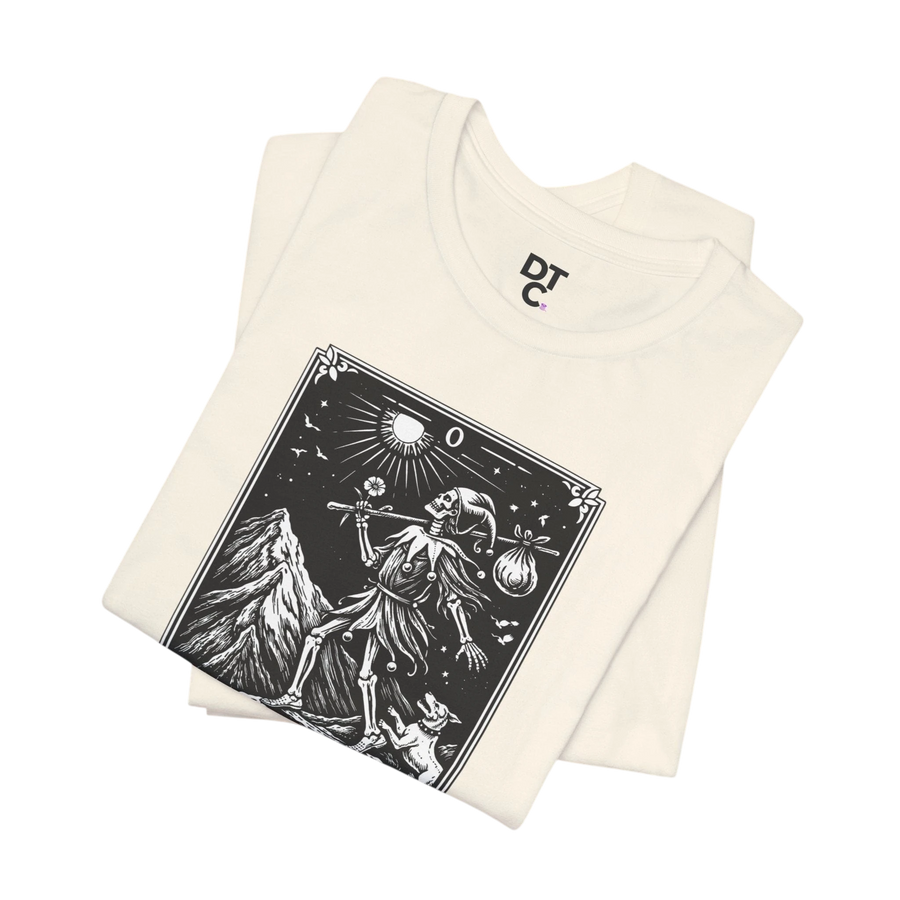 DailyThreadCo. cream cotton t-shirt with 'The Fool' tarot card graphic print in black and white, featuring a skeleton figure, mountain, sun, and dog.