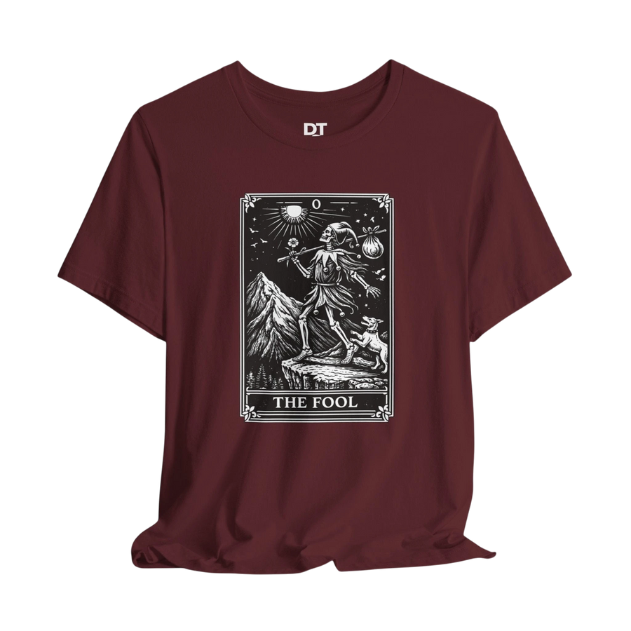 DailyThreadCo. maroon cotton t-shirt with 'The Fool' tarot card graphic print on front. Features skeleton figure, dog, mountains, and celestial symbols.
